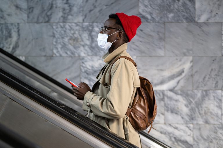 Afro-American traveler man stands on escalator in subway or railway station, wear face medical mask to protect yourself from contact with flu virus, pandemic covid-19